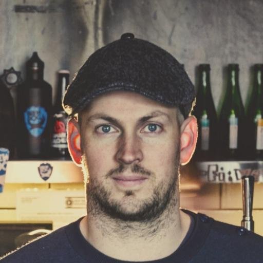 A close-up portrait of James Wattt with light eyes and a beard wearing a dark flat cap, and beer bottles on a shelf blurred in the background.