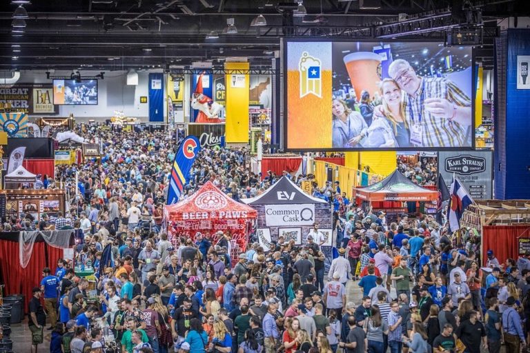 "A bustling crowd enjoys the Great American Beer Festival, featuring brewery booths like Beer Paws and Common Cask, with a large screen overhead displaying festive attendees enjoying craft beer."