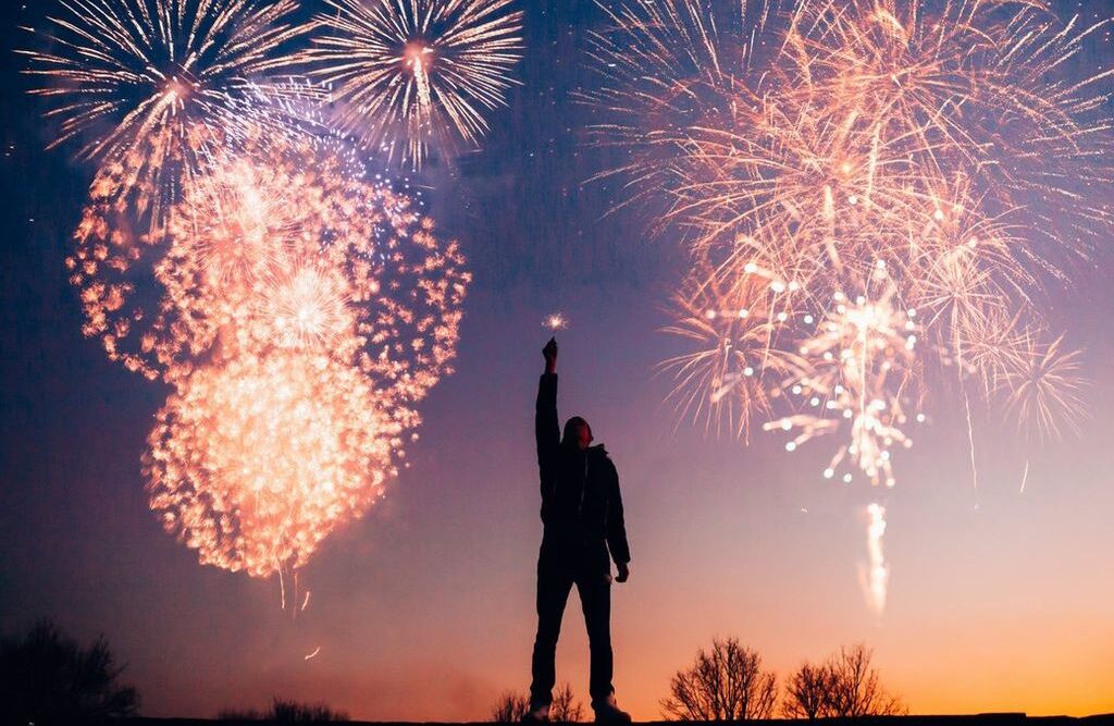 Silhouette of a person raising a hand toward a night sky filled with large, bright fireworks bursts.