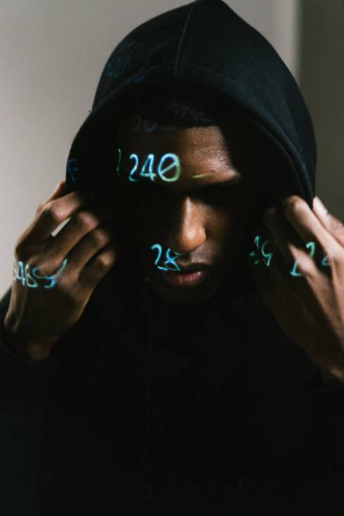 A man in a black hoodie with glowing digital numbers and data projected onto his face and hands, conveying themes of technology, data theft and computor hacking.
