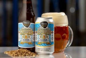 Tröegs Independent Brewing Oktoberfest Lager beer bottle, can, and full mug with frothy head, surrounded by barley grains, set against a blurred brewery background.