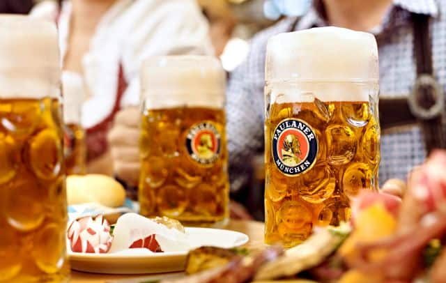 Paulaner Oktoberfest beer steins filled with golden lager on a table with traditional German food, capturing the festive atmosphere of an Oktoberfest celebration