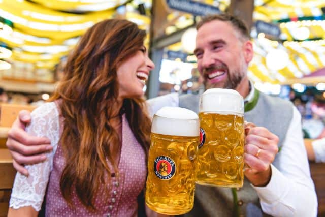A smiling man and woman, dressed in traditional Bavarian attire, toast with large Paulaner beer mugs at what appears to be a lively Oktoberfest celebration, with festive yellow and white decorations in the background)