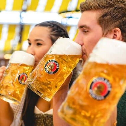 Three people are shown drinking large glass mugs of Paulaner beer at what appears to be an Oktoberfest celebration.