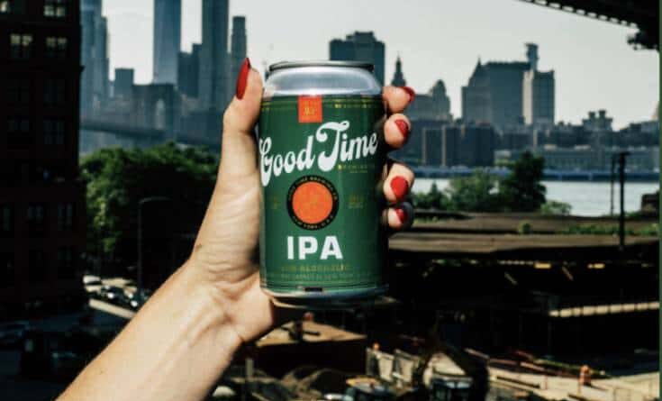 A hand with red fingernails holds up a green "Good Time IPA" beer can with an orange circle logo, in front of a blurry, sunlit city skyline with a river and bridge.