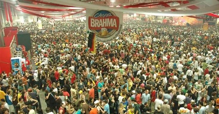 A lively indoor scene at Oktoberfest Blumenau in Brazil, showcasing a massive crowd, a large stage with a "Chopp BRAHMA" banner, and a vibrant festival atmosphere