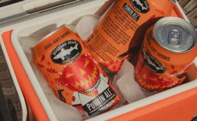 Dogfish Head Punkin Ale cans from the 2024 Art Series, featuring a distinct pumpkin-themed design, chill amongst ice in an orange and white cooler.