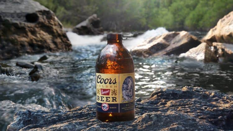 An amber bottle of Coors Golden Banquet Beer stands upright on a sunlit rock by a flowing river, surrounded by other rocks and green foliage in the background