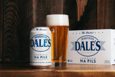 A can and glass of Oskar Blues Designated Dale's American NA Pils (non-alcoholic beer), alongside a six-pack box of the same product, all set against a dark, wooden background.