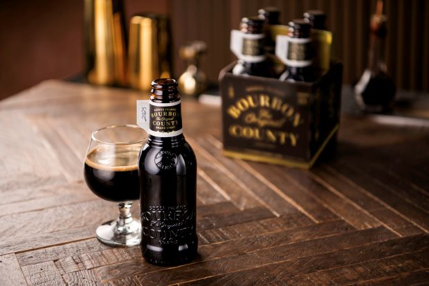 A single bottle of Goose Island Bourbon County stout beer next to a glass of the stout on a wooden table, with a four-pack carrier holding more bottles in the blurred background