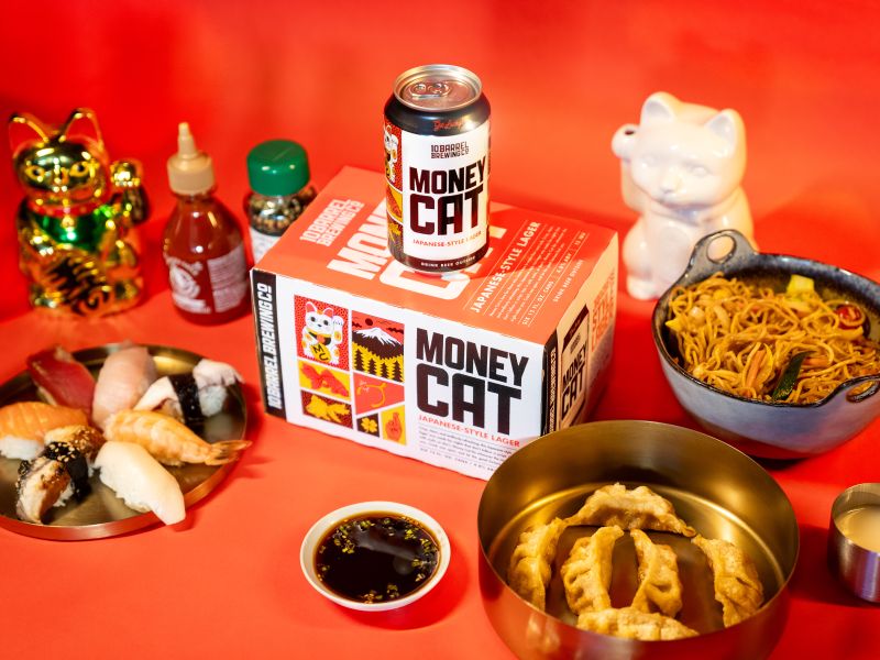 A spread of Japanese-style food and beer including sushi, noodles, dumplings, and a can and box of 10 Barrel Brewing Co. Money Cat Japanese-style lager, set against a red background with decorative cat figurine