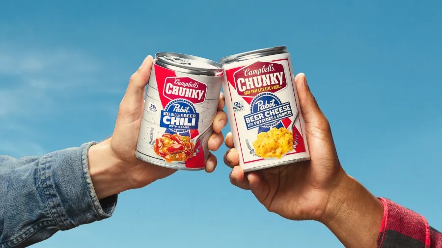 Two hands hold up cans of Campbell's Chunky Soup: one is "Beef, Bacon & Beer Chili with Beans" and the other is "Pabst Beer Cheese with Potatoes & Chorizo," set against a clear blue sky.