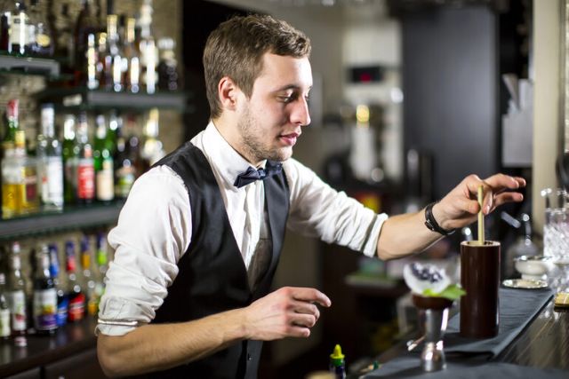 Bartender in a vest and bow tie preparing a drink behind a bar stocked with bottles