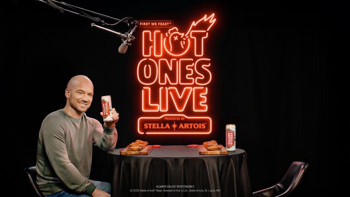 Sean Evans, host of "Hot Ones," at a "Hot Ones Live" event presented by Stella Artois, holding a Stella Artois beer can next to a table with hot wings