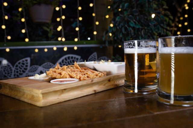 wo beer mugs and a wooden platter of fried food and dipping sauces on a table with festive string lights in the background