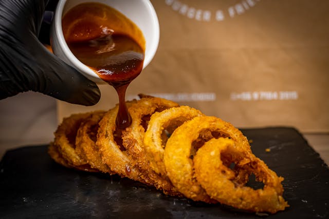 Crispy golden onion rings being drizzled with sauce on a dark serving plate, ready to ea