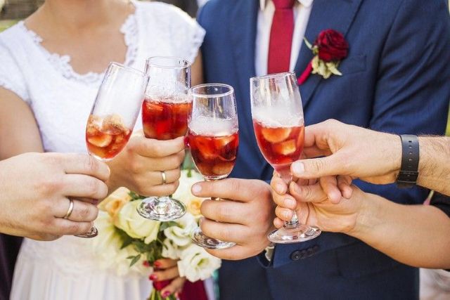 Wedding guests toast with glasses of red craft beer, celebrating a bride and groom dressed in white and navy blu