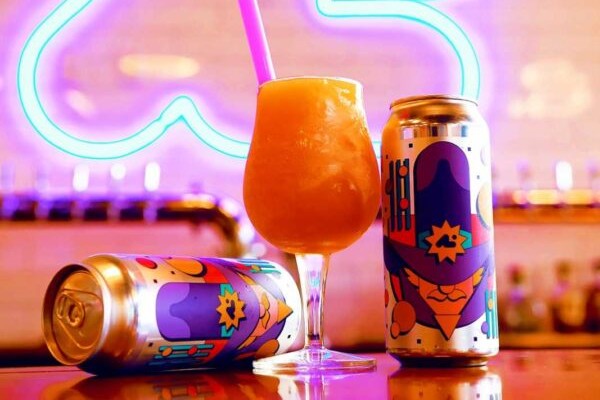 A vibrant, orange-hued Carrot Colada cocktail in a hurricane glass with a pink straw, flanked by two colorful cans from Aslin Beer Co., set against a background of blurred neon lights in a bar.