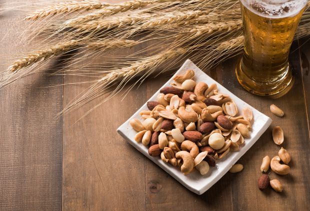 A close-up shot of a white square dish filled with mixed nuts (cashews, peanuts, almonds, macadamias) on a rustic wooden surface, alongside a frosted glass of beer and several stalks of barley
