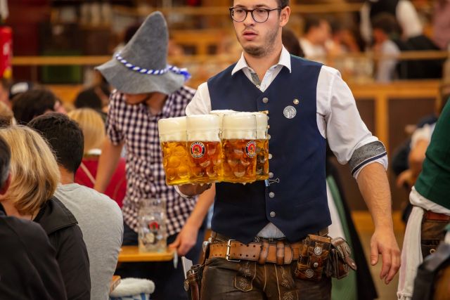 A man in traditional Bavarian attire, including lederhosen and a vest, carries a tray with several full beer steins through a crowded indoor setting, likely an Oktoberfest beer tent. He is wearing glasses and looking towards the right side of the frame. In the background, other people, some also in traditional clothing, are seated at tables.