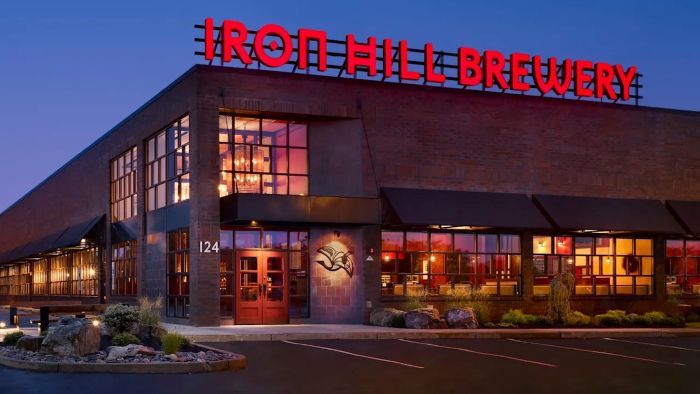 A nighttime exterior view of an Iron Hill Brewery & Restaurant location with its red neon sign illuminated, featuring large windows revealing the interior, and landscaping in the foreground