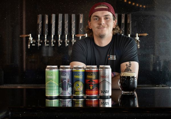 Pinnacle Brewing brewer behind bar with craft beer cans and a glass of Tori Japanese Rice Lager and beer taps in the background