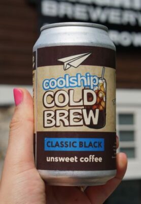 A hand holding a can of Coolship Cold Brew Classic Black unsweetened coffee. The can features a paper airplane logo and an illustration of an iced coffee drink
