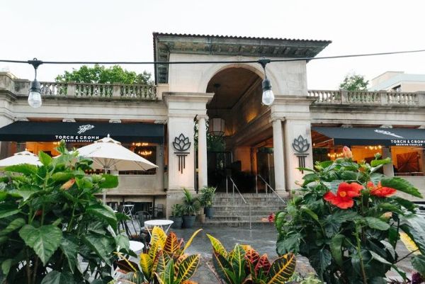 The exterior of Torch & Crown Brewing Company in Union Square, featuring a stone facade with arched entrances, black awnings over outdoor seating areas with white umbrellas and tables, and vibrant red and green plants in planters