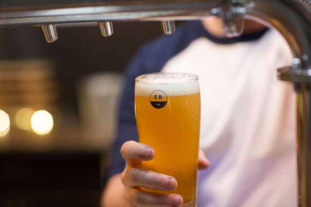 A hand holds a tall glass of golden beer with a white foamy head, positioned under a gleaming silver beer tap, likely in a bar or brewery setting. The beer glass has a small black circular logo with white text "EB" on it