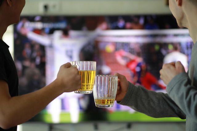 Two people clinking beer mugs while watching a soccer game on TV