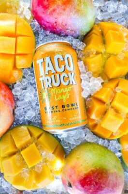 Dust Bowl Brewing Company's Taco Truck Mango Lager can surrounded by fresh mangoes and ice, highlighting the tropical flavor and refreshing nature of the craft beer