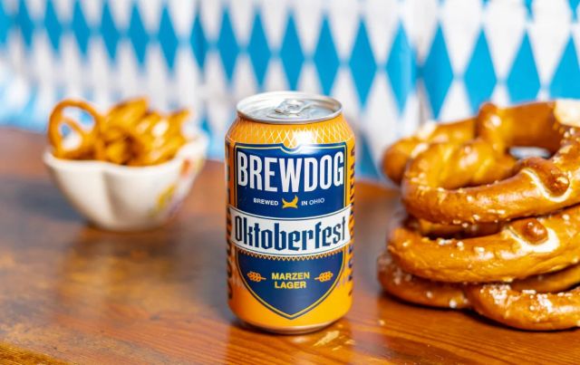 A can of BrewDog Oktoberfest Marzen Lager beer stands on a wooden surface, surrounded by pretzels. In the background, a blue and white checkered pattern evokes an Oktoberfest celebration.