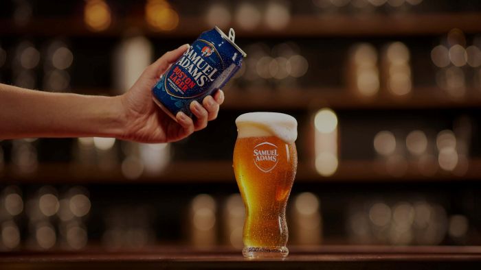 Close-up of Samuel Adams Boston Lager beer being poured from a can into a branded glass, showcasing the amber liquid and creamy foam.