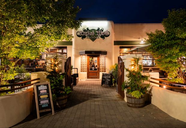 Exterior view of Wicked Weed Brewing pub in Asheville, NC at night, featuring illuminated signage, inviting entrance, and outdoor seating with lush greenery