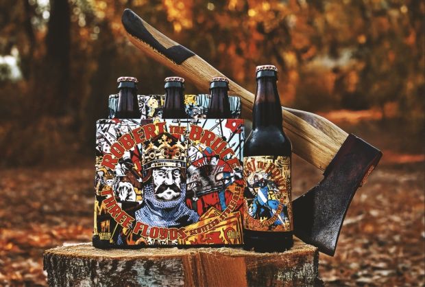 Three Floyds Robert the Bruce Scottish Style Ale six-pack and single bottle on a tree stump, with a large axe leaning against it, set in a rustic autumn backdrop