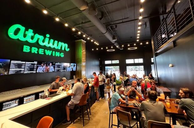 A wide shot of the interior of Atrium Brewing, a bustling brewery and taproom. The space has a high ceiling with exposed ductwork and string lights. A long bar with several people sitting at it is visible on the left. In the center and on the right, patrons are seated at wooden tables, eating and drinking.