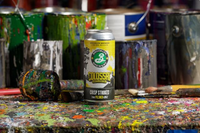 A close-up shot of a can of Brooklyn Brewery's Colossal Crisp Strokes Pilsner, a limited-release beer, placed on a paint-splattered workbench amidst paint cans and brushes.