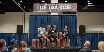 Three people are seated on stools, with a banner above them that reads "TAP TALK STAGE". The panelists are sitting on what look like metal beer kegs, and an audience is visible in the foreground. This event is part of the California Craft Beer Summit, which includes "Tap Talks" sessions