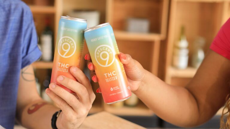wo people toasting with cans of Crescent 9 THC Seltzer, a tropical-flavored, alcohol-free beverage. The cans have a colorful gradient design and the text "Crescent 9 THC Seltzer" is visible on the front of each can. The background is a warm, indoor setting with shelves.