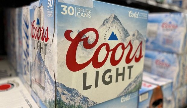 A large 30-pack case of Coors Light beer is seen on a shelf in a store. The case features a prominent red "Coors Light" logo and an image of a snowy mountain range with the phrase "Cold AS THE ROCKIES" printed on the box. Other beer cases, including Miller Lite, are partially visible in the background.
