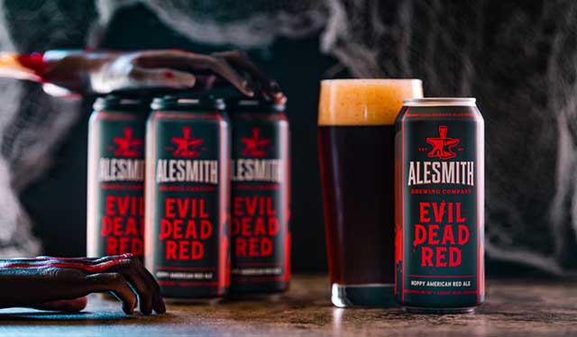 A promotional image featuring AleSmith Brewing Company's Evil Dead Red, a hoppy American red ale. The image showcases four cans of Evil Dead Red with bold red and black labeling, arranged in a row. A glass of the dark amber beer with a frothy head sits beside an open can. The background includes eerie, shadowy hands and a dark, textured setting, adding a spooky theme to the presentation.