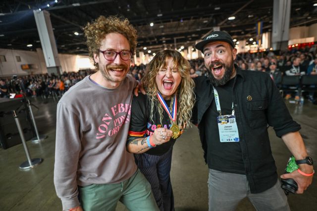 Three people, two men and one woman, are posing for a photo at an indoor event. The woman in the center is wearing a medal and has her mouth open in a shout or laugh. The two men on either side of her are also smiling and looking at the camera. The background is a large hall with a crowd of people.