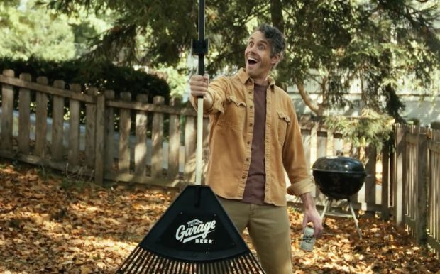 A man with an excited expression stands in a yard filled with autumn leaves, holding a leaf rake with a large plastic head that has "Garage Beer" printed on it. He holds a can of beer in his other hand.
