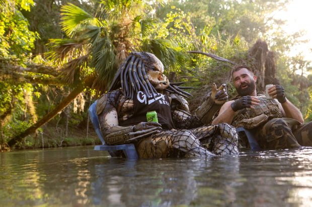 A person in a Predator costume and Jason Kelce in tactical gear sit in chairs in a river, with lush vegetation in the background. The person in the Predator costume is holding a Garage Beer can.