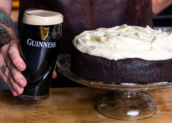 A close-up shot shows a rich, dark Guinness chocolate cake with a thick, white cream cheese topping on a glass cake stand, next to a hand holding a pint of Guinness stout.