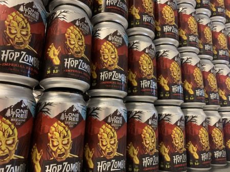 A large display of canned beer from Lone Tree Brewing, featuring a red and silver label with the brand name and the text "Hop Zombie" and "Imperial Red IPA." The label shows an illustration of a zombie's head made of a hop cone.