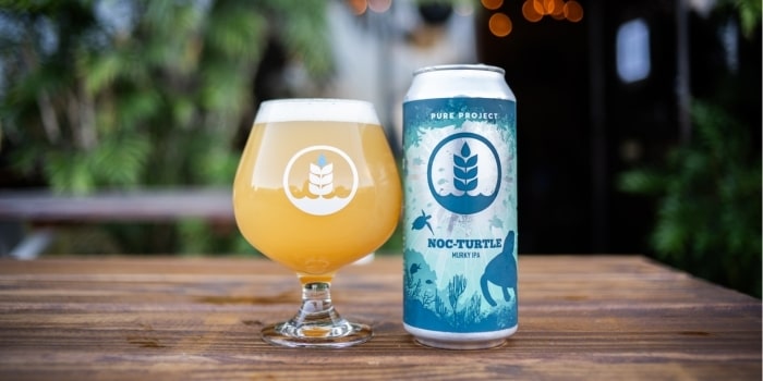 A can and a glass of Pure Project Noc-Turtle hazy IPA beer with a sea turtle design, sitting on a wooden table outdoors.