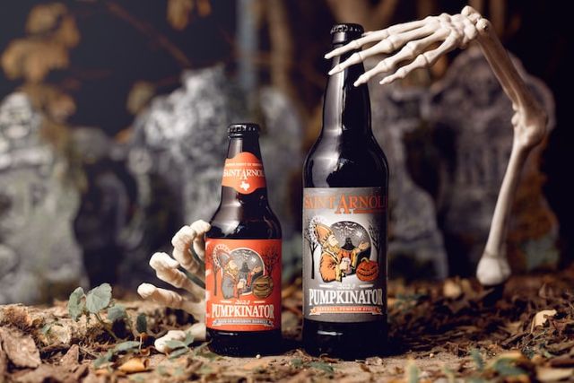 Saint Arnold Pumpkinator beer bottles in a Halloween-themed setting with skeleton hands and a spooky background