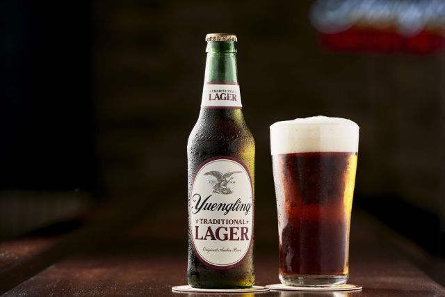 A bottle of Yuengling Traditional Lager next to a full glass of the same beer, with condensation on both containers, on a table against a black background.