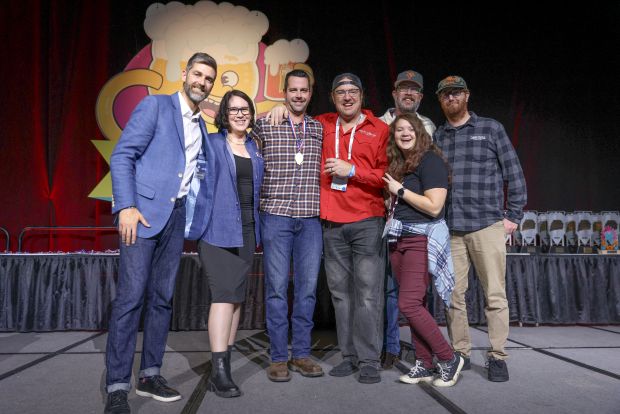 A group of six people, including four men and two women, are standing on a stage and smiling at the camera. In the background, there is a large, colorful sign featuring a cartoon beer mug. The people appear to be receiving Great American Beer Festival awards 